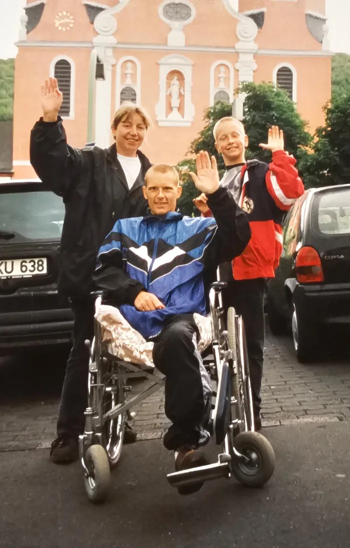 Dennie als jongen van twaalf, zwaaiend naast zijn vader Jos in de rolstoel en zijn moeder